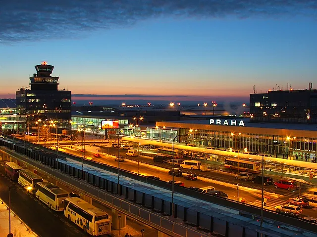 Václav Havel Airport Prague at dusk: the control tower and the terminal with the illuminated PRAHA sign, buses and cars on the forecourt under a dusky winter sky.