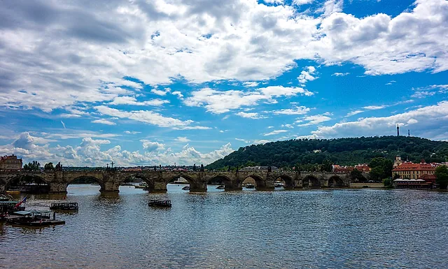 Charles Bridge's stone arches spanning the Vltava under a cloudy blue sky, with the wooded Petřín hill and its lookout tower rising behind the bridge.