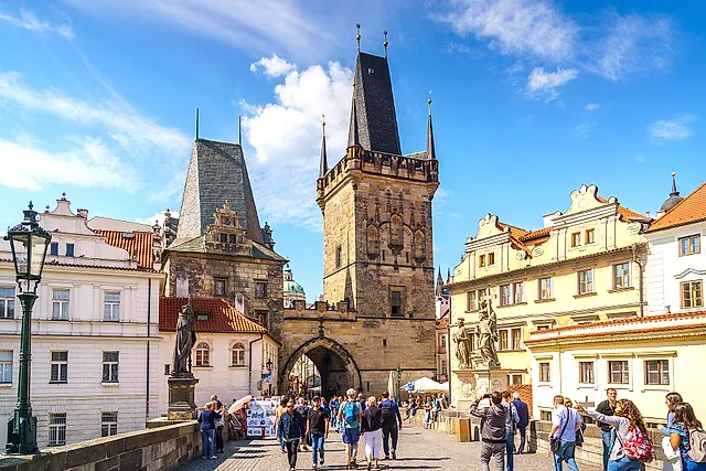 Gothic gate tower of Charles Bridge with a tall pointed roof and an arched passage below, tourists on the cobbled bridge and Baroque statues beside the tower.