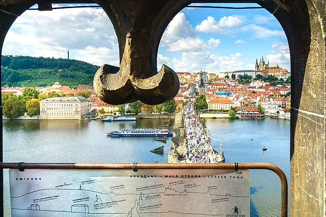 View from inside a stone bridge tower through a dark iron arch over the Vltava, the Charles Bridge and red-roofed houses, with castle spires at the right.