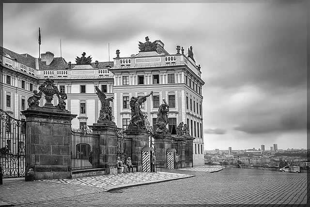 Black-and-white view of a Baroque palace behind wrought-iron gates and stone statue groups, with striped sentry boxes and a grey sky above the distant city.