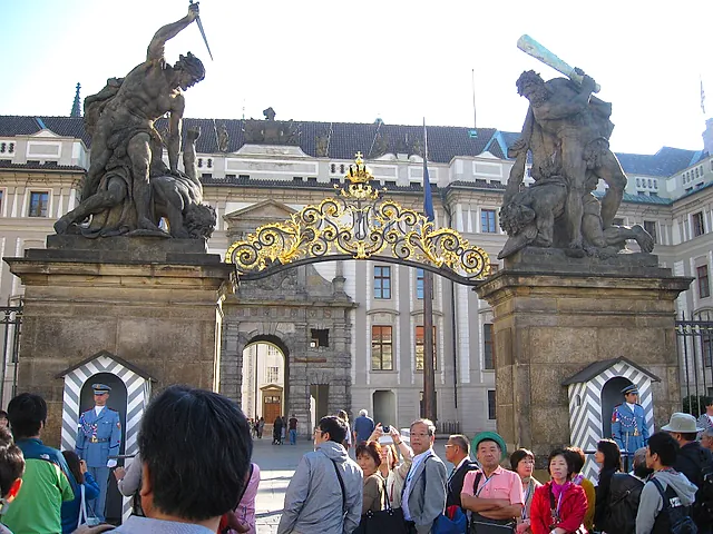 Ornate gilded wrought-iron gate under a Baroque arch, flanked by two stone statues of struggling giants, with tourists standing on the cobbles in front.