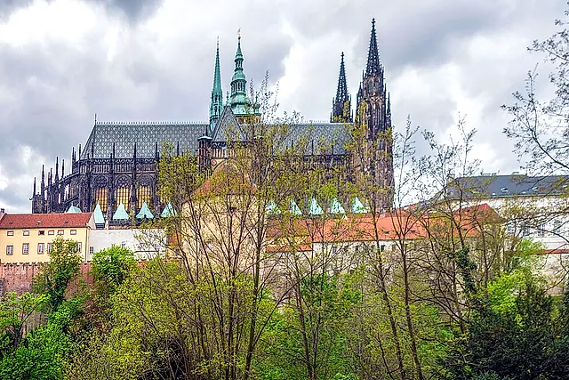 Wide view of St Vitus Cathedral's Gothic spires and roofs rising above trees and pale houses on the left, under a dramatic cloudy sky.