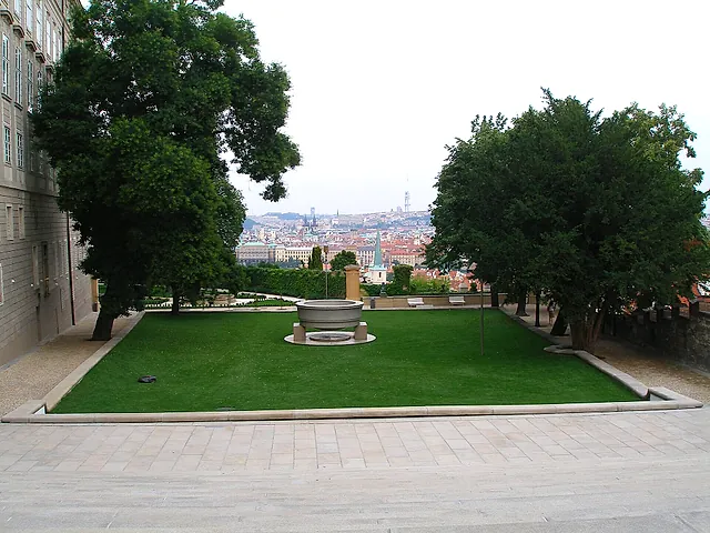 Lawn in a stone-edged garden with a round stone fountain basin in the middle, tall trees on both sides and a view of the city roofs beyond.