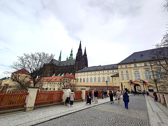 Cobbled road beside a wooden fence, with the cathedral spires behind a yellow Baroque palace with Czech flags, and visitors walking under a cloudy sky.