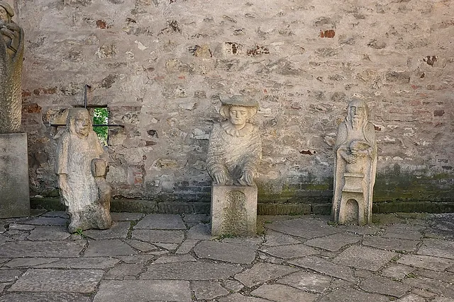 Weathered stone busts of figures standing against a rough stone wall with a small window, on an uneven flagstone pavement in a courtyard.