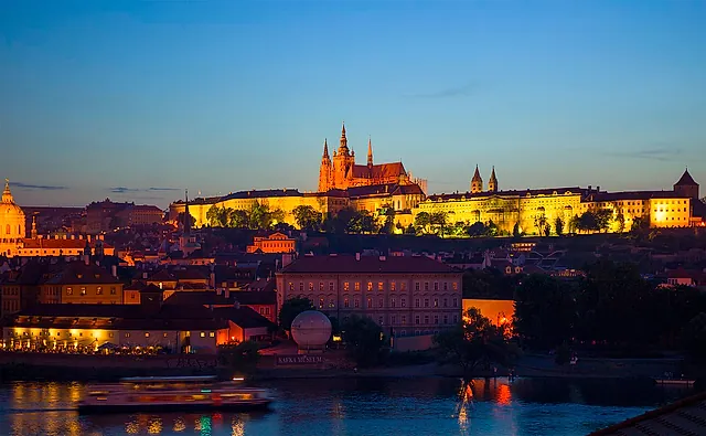 Prague Castle at dusk with floodlit cathedral spires and palace fronts above the dark rooftops, a tour boat on the river and a Kafka Museum sign below.