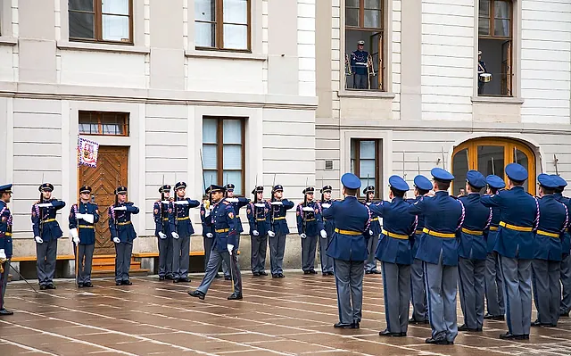 Ceremonial guards in dark-blue uniforms with white gloves stand in two lines on a wet courtyard before a cream palace with tall windows and an arched wooden door.