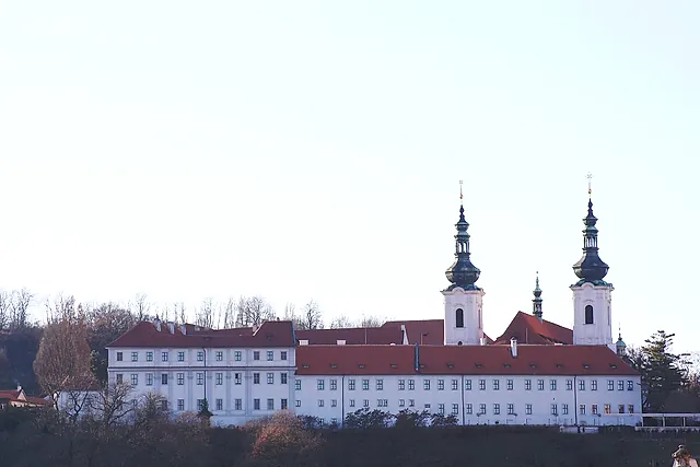 Strahov Monastery seen from below, its long white Baroque facade with rows of windows, red roofs and two onion-domed towers against a pale sky.