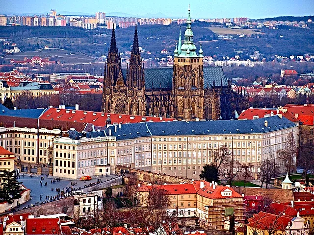 Prague Castle from Petřín hill: the dark Gothic cathedral spires and long palace range rise above the orange rooftops of the Lesser Town in soft light.