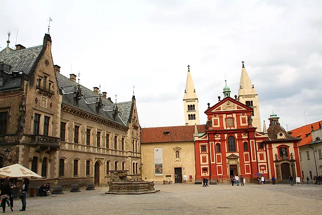 Square at Prague Castle with the red-and-white Baroque facade of St George's Basilica and its two pale towers, an ornate gabled building on the left and visitors on the cobbles.