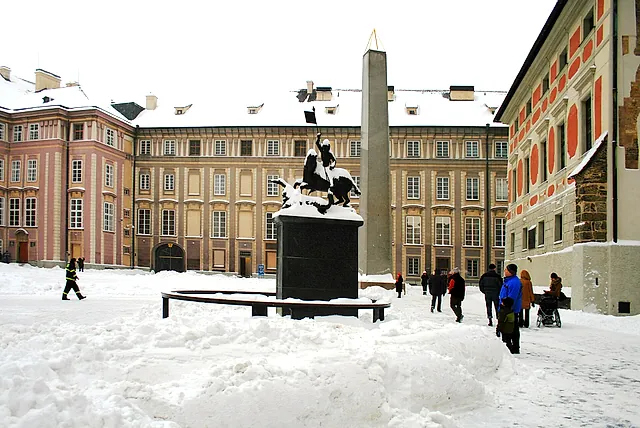 Snowy courtyard with a black equestrian statue of St George on a stone plinth, a tall grey obelisk behind it and Baroque palace facades around the square.