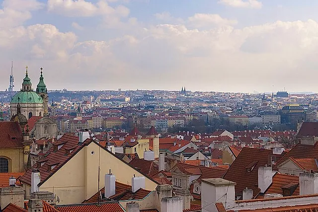 Panoramic view from the castle over red-tiled roofs and a green copper dome, with the city stretching to modern towers on the horizon under a pale sky.