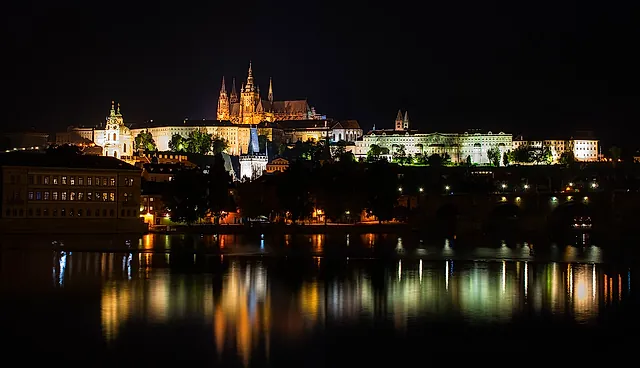 Prague Castle and the riverfront at night: the cathedral towers and palace facades are floodlit above dark rooftops, with the water reflecting the lights.
