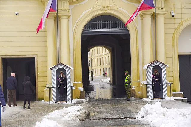 The arched gateway of Prague Castle with two striped sentry boxes and guards in dark uniform beside the Czech flags, with snow on the cobbles.