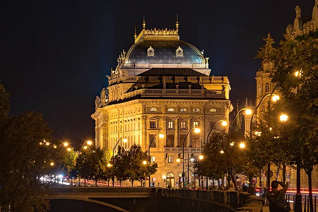 The National Theatre in Prague at night, its neo-Renaissance facade lit with warm light, a domed roof above and trees and lamp posts along the embankment.