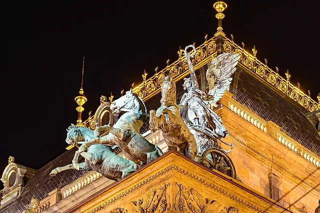 Gilded ornaments and a bronze horse-drawn chariot group on the roof of the National Theatre at night, lit from below against a dark sky.