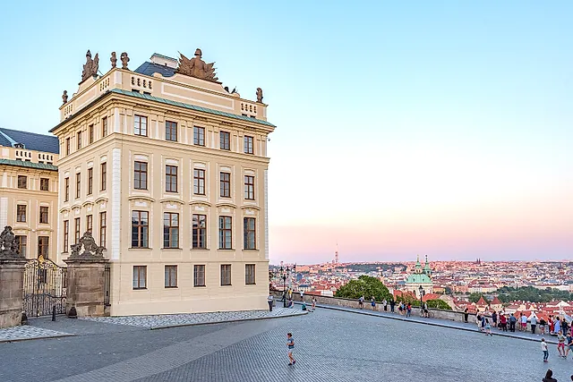 A yellow Baroque palace corner at Prague Castle with a sculpted roofline, and a paved terrace where visitors walk by the balustrade, with the city and a church dome beyond.