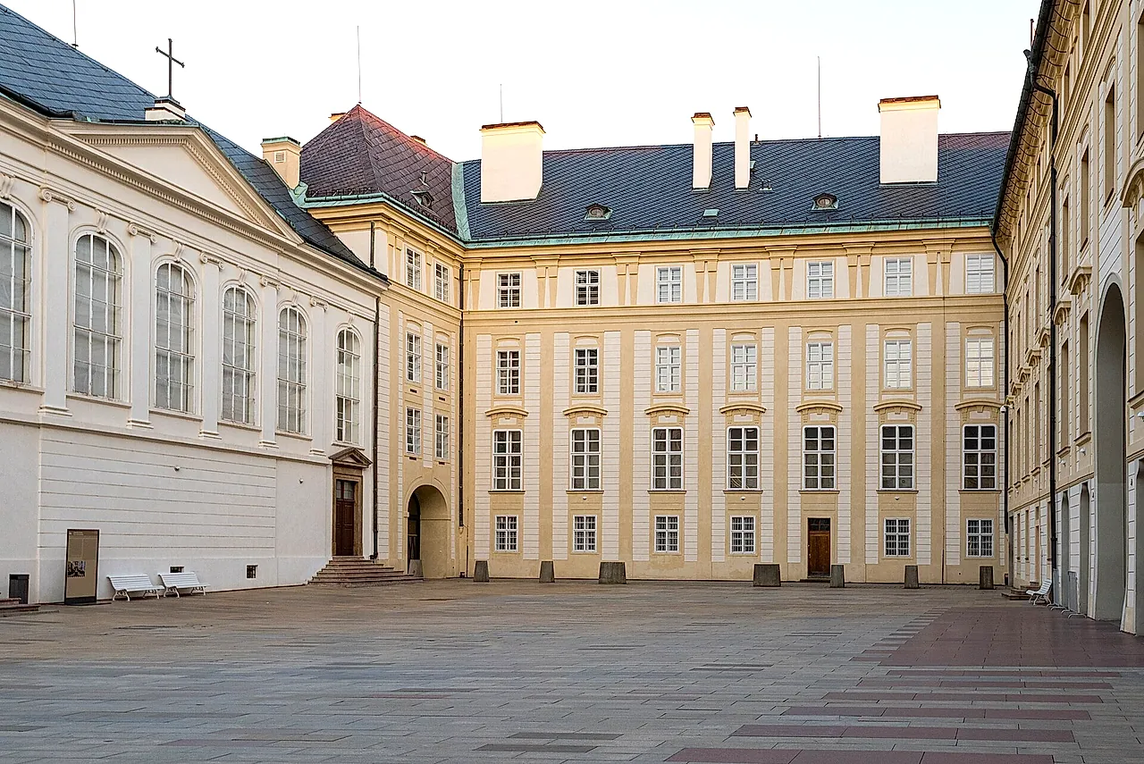 The Second Courtyard of Prague Castle, a wide paved square lined with cream Baroque facades, tall windows and a few white benches, under a pale sky.
