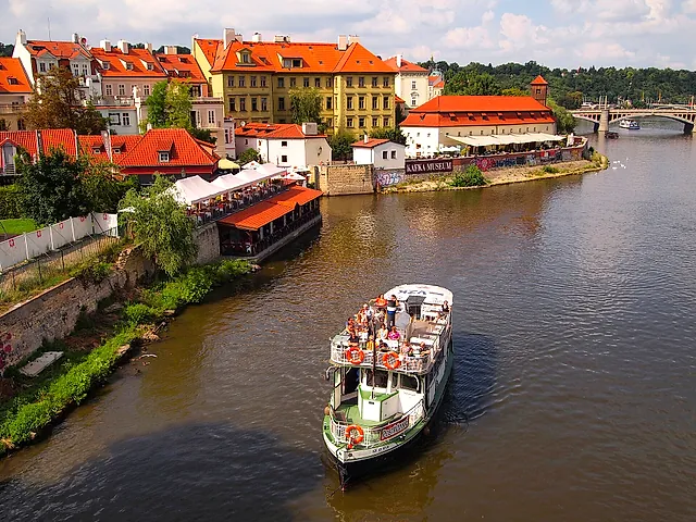 A white tourist boat with green trim and passengers on its open deck sails on the brown Vltava, with red-roofed houses and a bridge behind it.