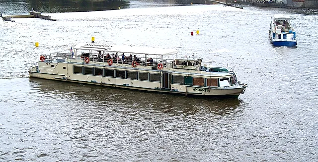 A long white cruise boat, its name on the hull, crosses the Vltava under grey skies, with passengers on the upper deck and a small blue boat behind.