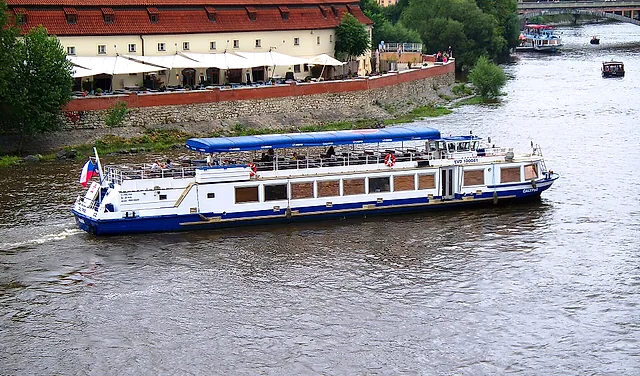 A white two-deck tour boat with a blue awning sails along the Vltava past a cream riverside terrace with white umbrellas, and another boat in the distance.