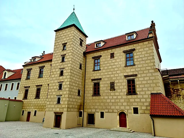 A castle building with a square corner tower under a green copper cap, walls with a stone-block pattern and a red-tiled roof, above an empty paved courtyard under a pale blue sky.