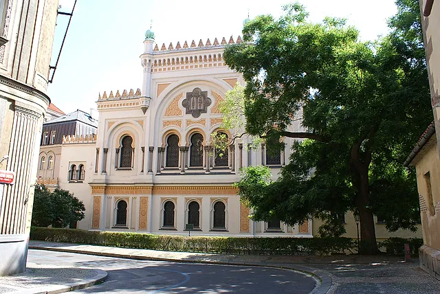 Facade of the Spanish Synagogue in Prague, a cream building with arched windows and a crenellated roofline, partly hidden by a large tree on a quiet street.