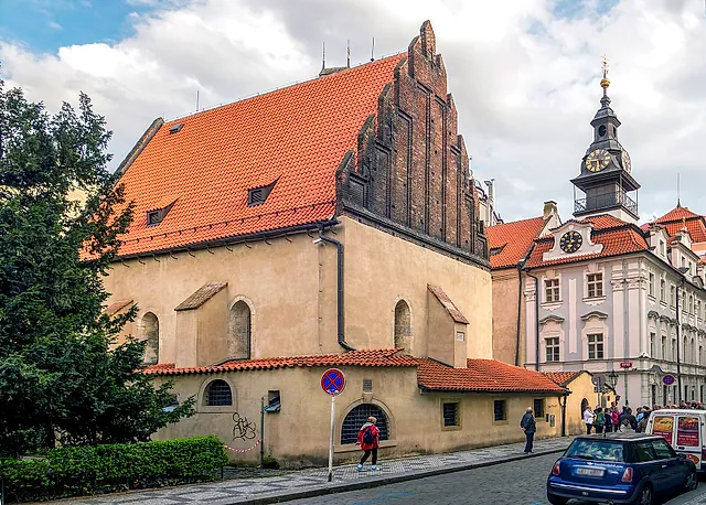 Old-New Synagogue in Prague, a Gothic hall with a stepped gable and steep red-tiled roof, beside a pink building with a clock tower under a cloudy sky.