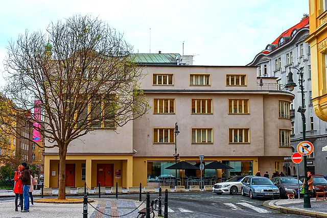 Pale building with rows of tall windows on a city square, a bare tree in front, a few people and parked cars nearby under a pale sky.