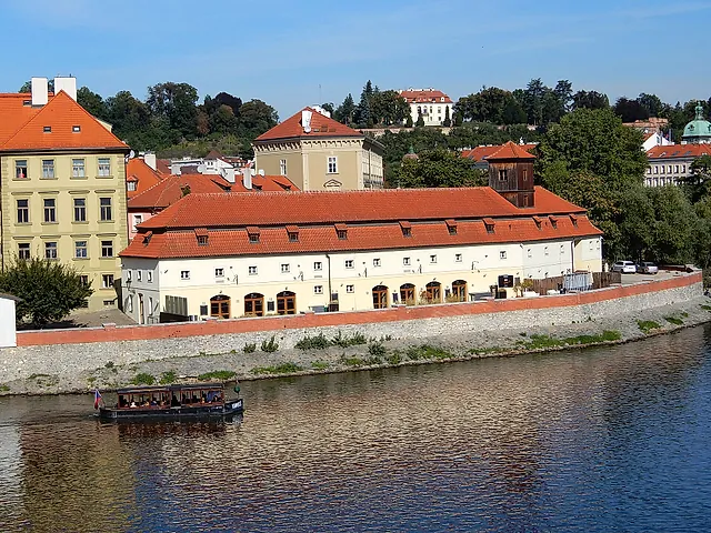 Long cream-coloured museum building with a red-tiled roof and arched windows on the Vltava embankment, with a tour boat on the water and trees behind.