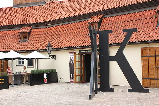 Tall black steel letter K standing in a paved courtyard in front of a cream building with a red-tiled roof, with a café terrace under white umbrellas on the left.