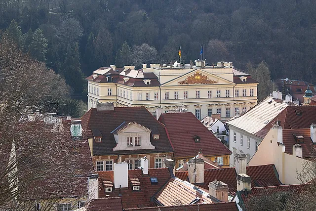 Pale yellow Baroque palace with a sculpted central pediment and two flags on its roof, seen above red roofs and dark wooded slopes in winter light.
