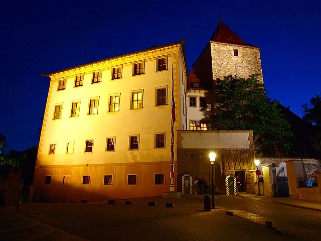 Cream-coloured palace at night with rows of lit windows, a red-roofed stone tower behind it, a flagpole and a lamp post on the cobbled square.