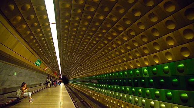 Platform of Malostranská station on Prague Metro line A, with a curved wall of round panels lit in green and a few passengers waiting along the platform.