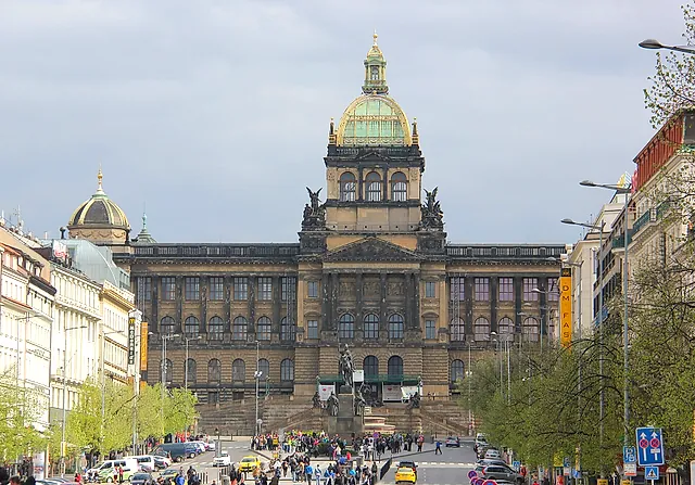 The National Museum in Prague, a large building with a green copper dome, seen under grey skies with traffic on the avenue and small figures in the distance.