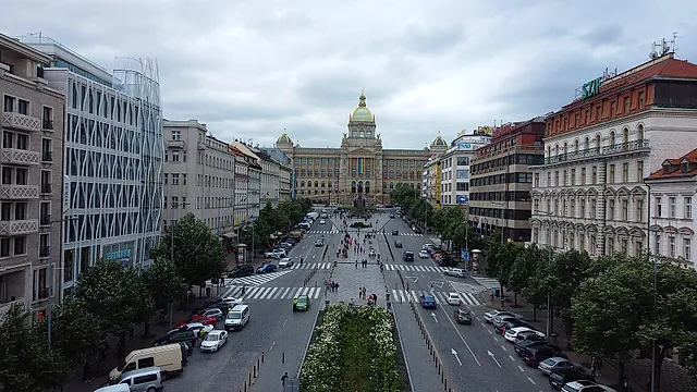 Wenceslas Square seen from above, a long boulevard with parked vans, trees and pedestrians, leading to the green dome of the National Museum far away under clouds.
