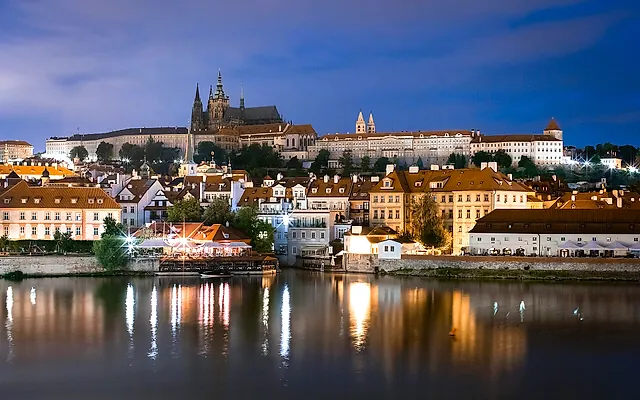 Prague Castle and the riverfront below it lit up at dusk, with St Vitus Cathedral's towers above the illuminated houses and their reflections in the calm Vltava.