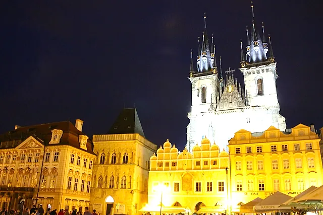 Old Town Square at night, with the twin spires of the Týn Church rising above floodlit Gothic and Baroque houses and a few small figures in the lower corners.