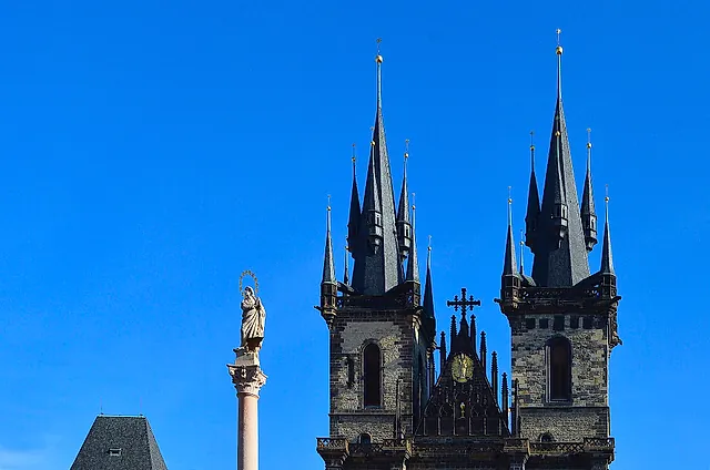 Dark Gothic spires of the Týn Church against a deep blue sky, with a column topped by a statue at the lower left and rooftops below.