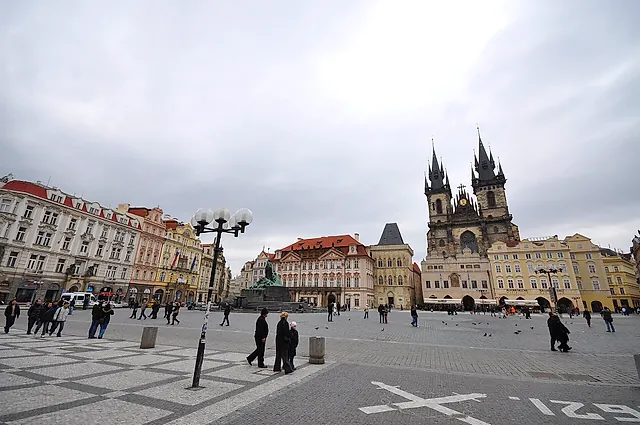 Old Town Square under grey daylight, with the Týn Church towers on the right and pastel facades around the square, and a few people walking across the paved surface.