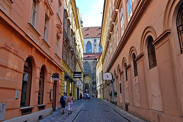 A narrow cobbled lane in the Old Town, with orange and pale-yellow house fronts, arched doorways and a Gothic church at the end, with a few distant pedestrians.