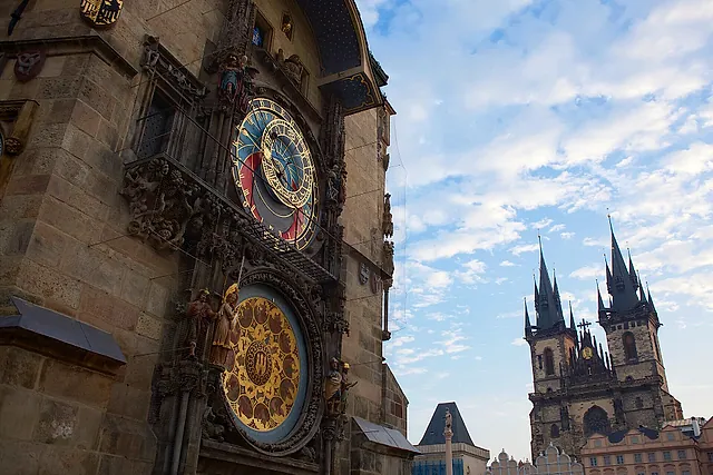 The astronomical clock dial on the Old Town Hall tower, with the twin Gothic spires of Týn Church rising against a cloudy blue sky behind it.