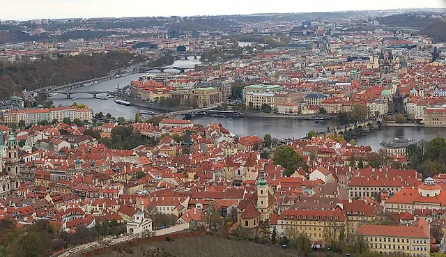 Panoramic view over Prague's red-roofed Old Town, with the Vltava winding through the city, its bridges crossing the river and church spires above the rooftops.