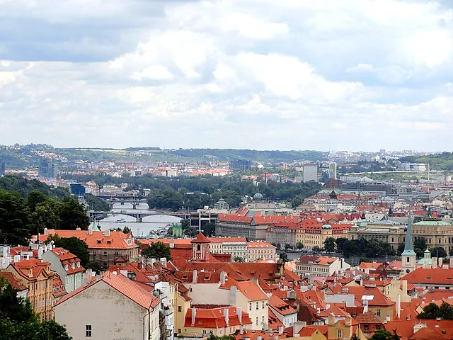 Panoramic view over Prague's red-tiled rooftops toward the Vltava and its bridges, with green hills and modern towers on the far bank under a cloudy sky.