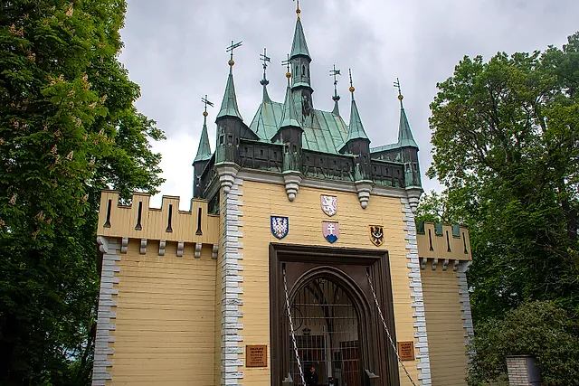 Entrance gate of the Mirror Maze on Petřín hill: a pale yellow turreted building with green copper roofs, a wooden door and heraldic shields above the arch.