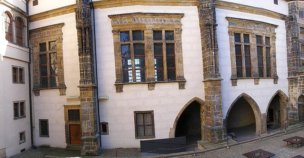 Una parete intonacata di bianco dell’Antico Palazzo Reale al Castello di Praga, con finestre incorniciate in pietra, due aperture ad arco acuto e contrafforti angolari in pietra squadrata sopra un cortile lastricato.