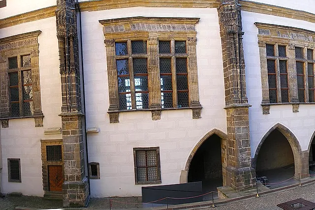 A whitewashed wall of the Old Royal Palace at Prague Castle, with stone-framed windows, two pointed-arch openings and corner buttresses of dressed stone above a paved yard.