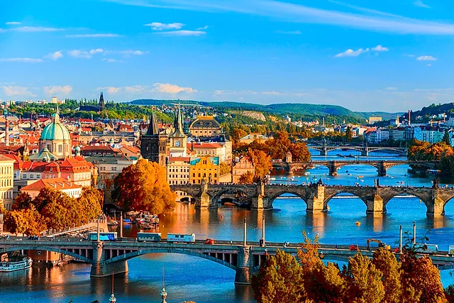 Panoramic view over Prague's rooftops in autumn, with a green-domed church, orange trees and arched bridges crossing the river under a blue sky.