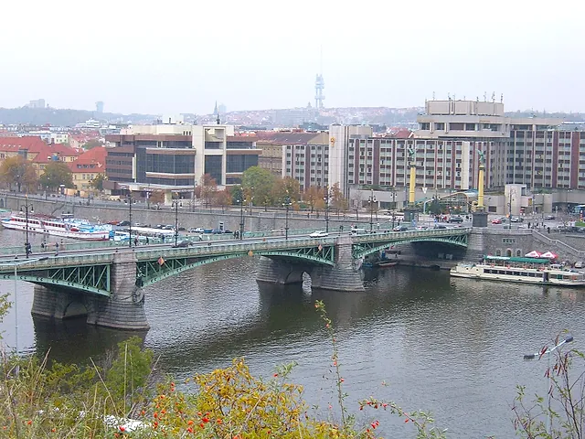 View from Letná Park over the Čech Bridge, a green steel bridge spanning the Vltava, with white boats moored on the bank and office blocks behind.